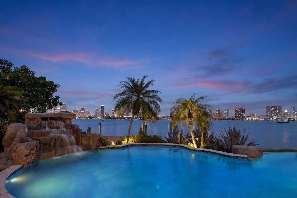 This stunning twilight view captures a custom swimming pool featuring a naturalistic rock waterfall feature on the left. The pool overlooks a calm bay with a panoramic skyline of a city visible in the distance under a soft, colorful sunset sky. Two tall palm trees frame the center of the composition, creating a serene and luxurious waterfront atmosphere.