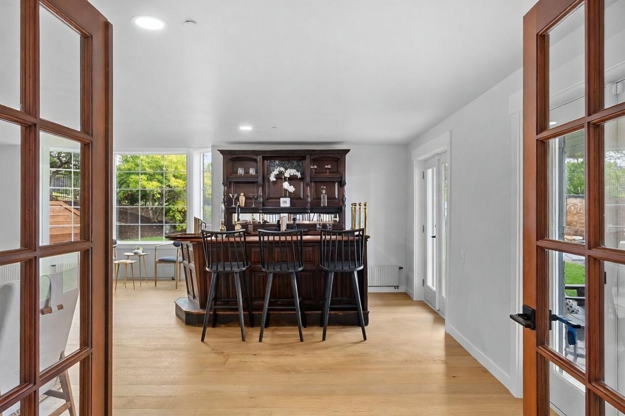 This interior view is captured from a doorway looking into a home bar area featuring a dark wood bar with three black spindle-back stools. The room is brightened by large windows that look out onto a green yard, and the space is finished with light wood flooring. The perspective creates a welcoming transition from the foreground into the central entertainment area.