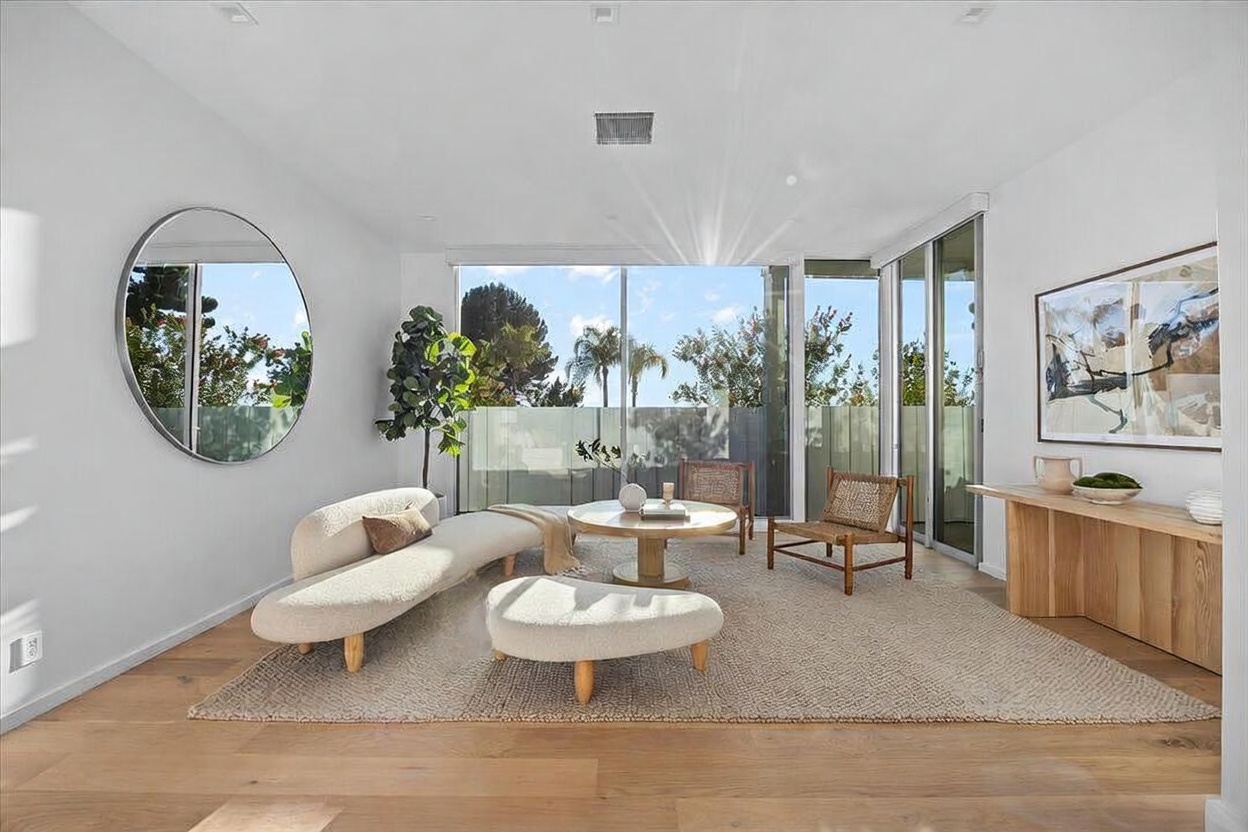 This bright and airy living room features a contemporary aesthetic with light wood flooring and a neutral color palette. The space is furnished with a curved sofa, a matching ottoman, and two woven accent chairs arranged around a circular coffee table, all set atop a large textured area rug. Large sliding glass doors provide an expansive view of the outdoor area, while a large circular mirror on the wall enhances the sense of space and light.