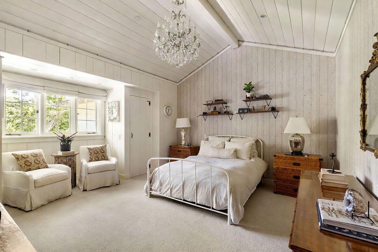 This bright and airy bedroom features a vaulted ceiling with exposed beams and a central crystal chandelier, creating an elegant, cottage-inspired aesthetic. The room is furnished with a white metal bed frame, two wicker-style nightstands, and a cozy seating area by the window. Vertical wood paneling on the walls and soft neutral carpeting contribute to a warm, inviting atmosphere.