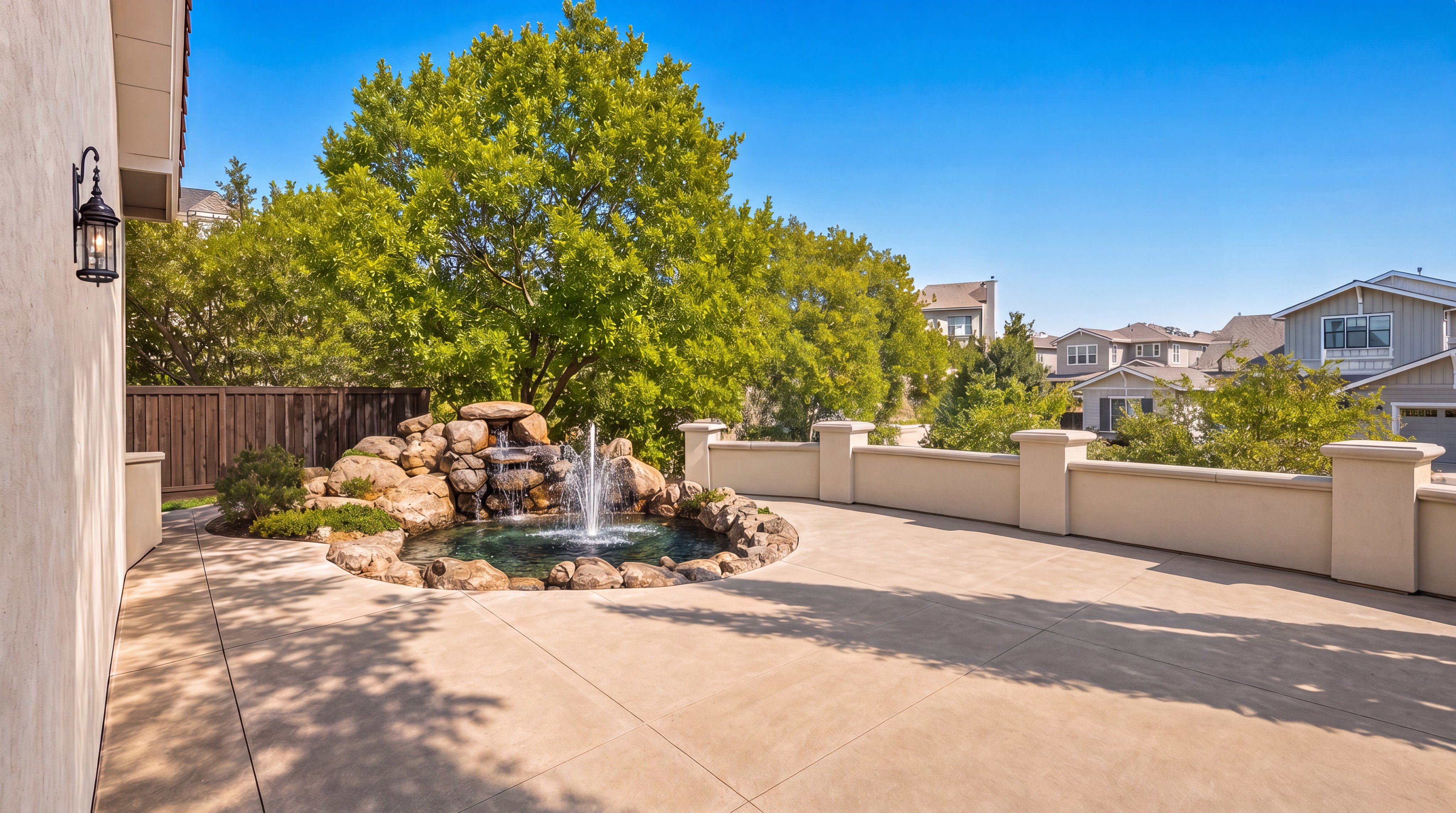 This spacious, modern patio features a decorative rock waterfall fountain, creating a peaceful and upscale outdoor living area. The space is defined by clean, light-colored concrete flooring and a solid perimeter wall, offering a private setting with lush tree views. It serves as an excellent entertainment space that seamlessly blends luxury landscaping with functional outdoor design.