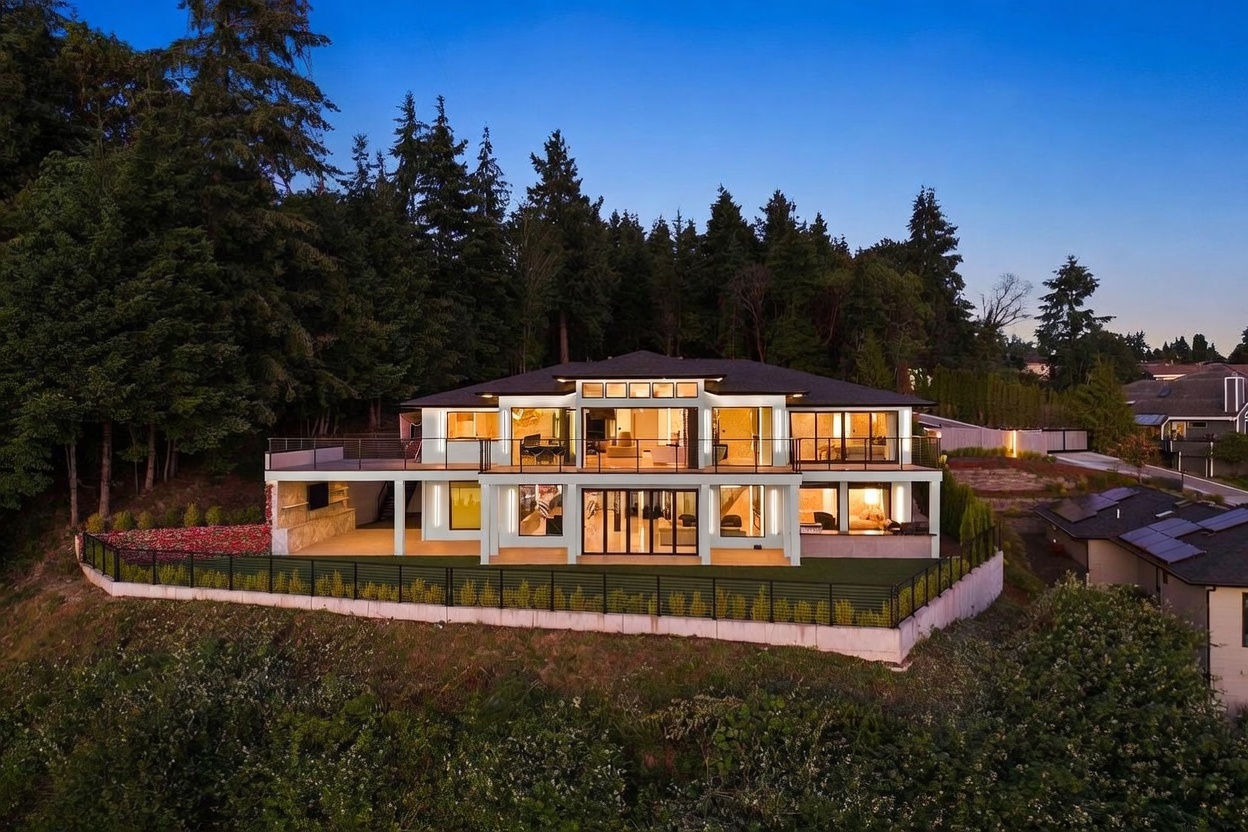 This contemporary two-story residence is captured from a rear perspective at dusk, showcasing a symmetrical design with extensive glass walls and expansive balconies. The home features a clean, modern aesthetic with a flat roofline, integrated outdoor lighting, and a terraced backyard enclosed by a dark metal fence. The elevated position provides a commanding view of the surrounding landscape, emphasizing the home's seamless indoor-outdoor living capabilities.