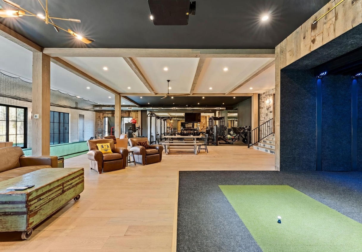 This expansive, multi-functional recreational space features a dedicated golf simulator area in the foreground, transitioning into a comprehensive home gym equipped with various weight machines and fitness equipment. The room showcases a rustic-industrial aesthetic with exposed wooden beams, light wood flooring, and a dark ceiling, creating a sophisticated yet rugged atmosphere. A comfortable lounge area with leather armchairs and a vintage-style trunk coffee table provides a relaxed space to unwind, while large windows and netting suggest a versatile layout suitable for both exercise and entertainment.