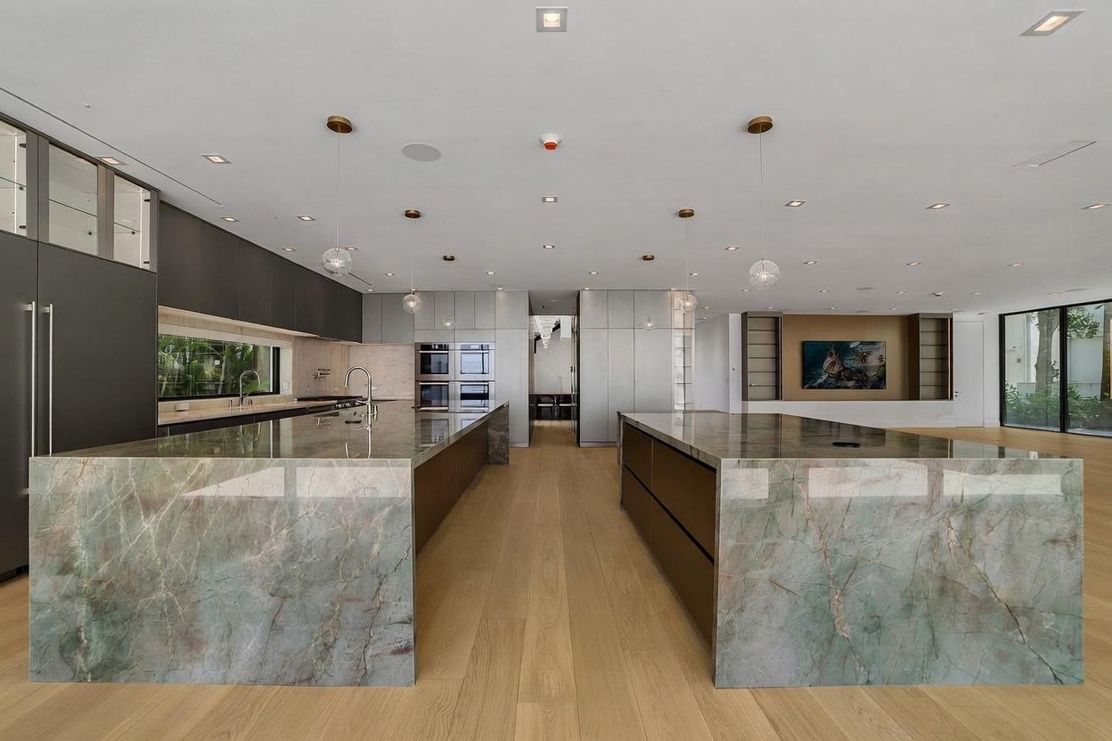 This expansive, modern kitchen features dual large islands with striking green-veined stone surfaces and dark cabinetry. The space is illuminated by minimalist pendant lights and recessed ceiling lighting, set against light wood flooring that runs throughout the open-concept area. A sleek, integrated appliance wall and a view into the adjacent living space create a sophisticated, high-end atmosphere.