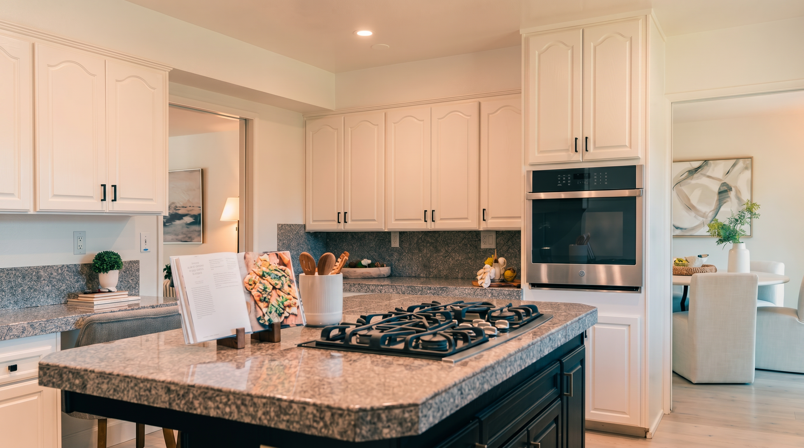 This bright and inviting kitchen features white cabinetry paired with a dark-toned central island topped with speckled stone countertops. A built-in wall oven is integrated into the cabinetry, while a gas cooktop is conveniently positioned on the island. The space is open and airy, with views leading into an adjacent dining area and a secondary room, creating a cohesive and functional layout.