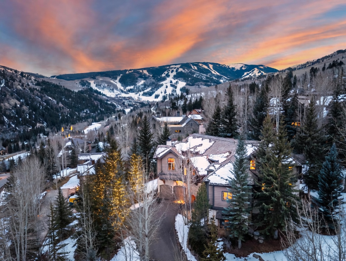 This elevated aerial perspective captures a luxurious mountain residence nestled within a snow-dusted, forested landscape at dusk. The home features a multi-gabled roofline, warm interior lighting glowing through the windows, and a paved driveway, all set against a dramatic, vibrant sunset over the distant ski slopes. The composition highlights the property's secluded, high-altitude setting and its integration into the natural alpine environment.