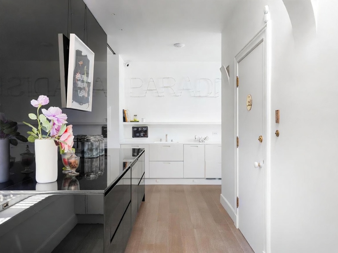 This interior view captures a sleek, modern hallway leading toward a bright, minimalist kitchen area. The space features light wood flooring, a high-gloss black surface on the left, and a white door on the right, creating a sharp contrast in textures. The far wall is adorned with large, white dimensional lettering that reads 'PARADISE', adding a contemporary artistic touch to the clean, gallery-like aesthetic.
