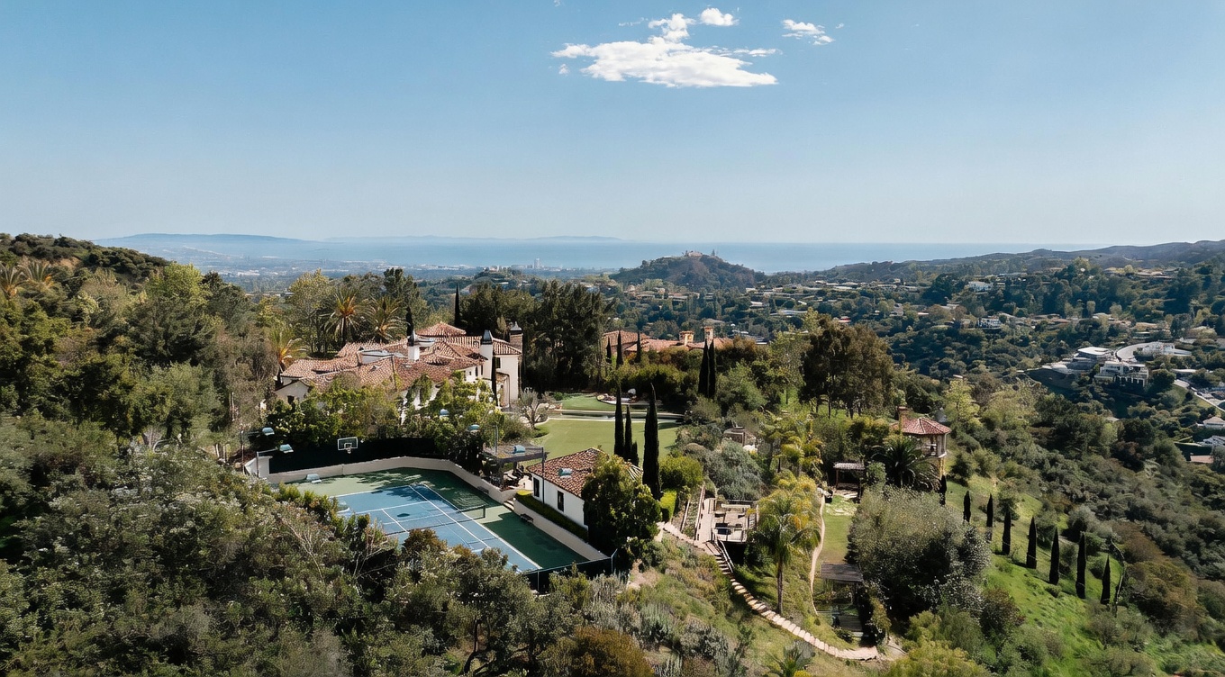 This high-angle aerial view captures a sprawling Mediterranean-style estate nestled within a lush, hilly landscape. The property features a private tennis court, a manicured lawn, and a multi-winged residence with a terracotta-tiled roof, all set against a backdrop of rolling hills and a distant ocean horizon. The perspective emphasizes the expansive nature of the grounds and the secluded, scenic setting of the home.