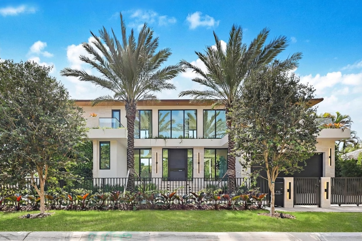 This modern two-story residence features a clean, symmetrical facade with a prominent central entryway and large windows that allow for abundant natural light. The exterior is accented by two tall palm trees framing the structure, a manicured lawn, and a sleek black metal fence with a matching gate. The overall aesthetic is contemporary and sophisticated, showcasing a well-maintained property with a clear view of the front elevation.