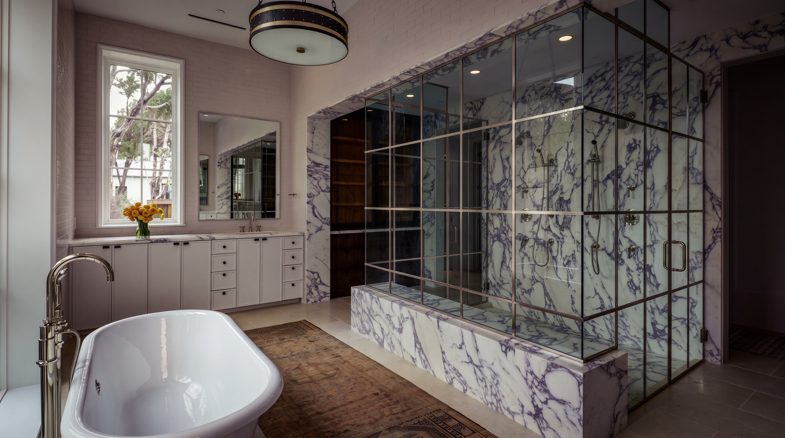 This luxurious primary bathroom features a freestanding white soaking tub positioned on a vintage-style rug, complemented by a large walk-in shower enclosed in a grid-patterned metal frame. The shower area is finished with striking patterned stone walls, while a long vanity with white cabinetry and a stone countertop provides ample storage. A large circular pendant light hangs from the ceiling, and a tall window allows natural light to brighten the space, creating an elegant and sophisticated atmosphere.