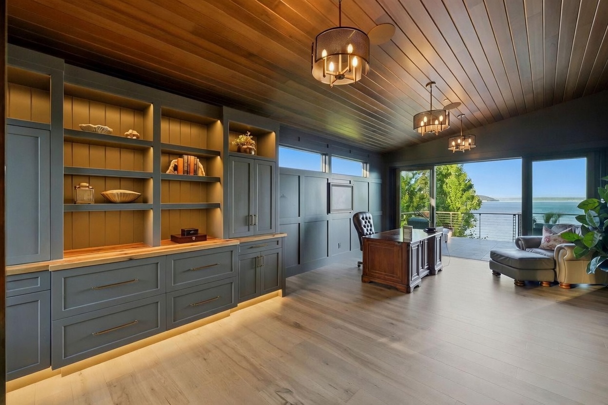 This sophisticated home office features custom dark-toned built-in cabinetry with integrated under-shelf lighting and a natural wood countertop. The room is anchored by a traditional wooden desk and a leather chair, complemented by a cozy seating area near the large sliding glass doors that provide expansive water views. The space is finished with a warm wood-paneled ceiling and modern drum-style pendant lighting, creating a refined and tranquil workspace.