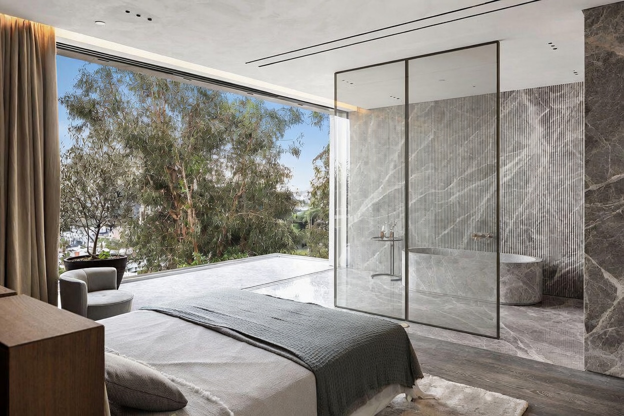This luxurious primary bedroom features a seamless transition between the sleeping area and an open-concept ensuite bathroom, separated by a sleek glass partition. The room is defined by its floor-to-ceiling windows that offer expansive, elevated views of lush greenery, creating a serene and sophisticated atmosphere. High-end finishes, including marble flooring and textured wall paneling, contribute to the modern, minimalist aesthetic of this high-end residence.