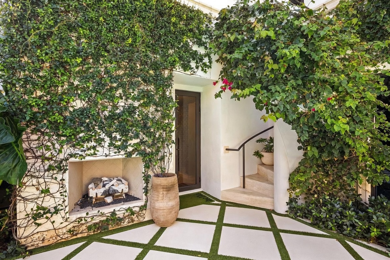 This inviting outdoor entryway features a unique stone-clad fireplace built into a wall covered in lush climbing vines. A paved patio with geometric grass inlays leads to a dark-framed glass door, while a curved staircase with a simple metal railing ascends to the right. The space is framed by vibrant greenery and bougainvillea, creating a serene and private atmosphere.