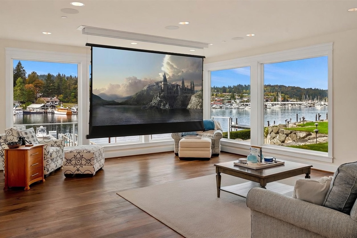 This spacious living room features a retractable projector screen displaying a scenic landscape, positioned centrally to take advantage of the expansive water views. The room is framed by large windows that offer panoramic vistas of the harbor and surrounding hills, filling the space with natural light. The interior is furnished with comfortable seating, including a patterned armchair and a plush sofa, set upon a neutral area rug over dark wood flooring.
