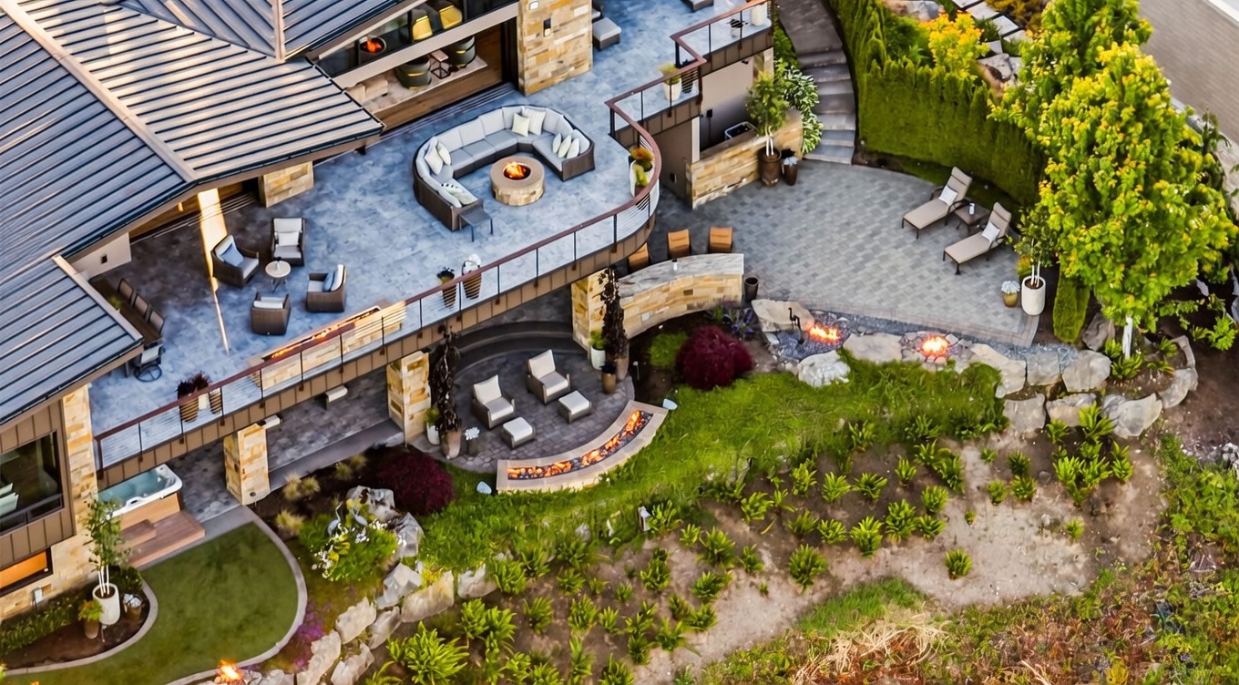 This high-angle aerial view captures a luxurious multi-level outdoor living space, featuring a spacious upper deck with a curved seating area and a fire pit. Below, a stone-paved patio offers additional lounge seating and a curved fire feature, seamlessly integrating with the landscaped hillside. The property showcases high-end finishes, including stone masonry and modern railings, creating an inviting and sophisticated atmosphere for outdoor entertaining.