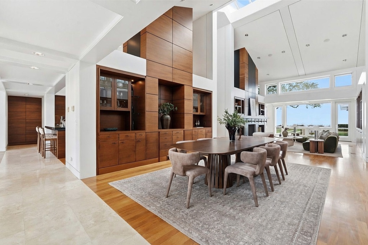 This expansive, open-concept dining area features a large wooden table surrounded by six upholstered chairs, set upon a textured area rug. A prominent wood-paneled wall unit with integrated shelving and cabinetry serves as a focal point, while the room flows seamlessly into a bright living space with tall windows overlooking the outdoors. The high, vaulted ceiling and light-toned wood flooring create an airy, sophisticated atmosphere suitable for formal entertaining.