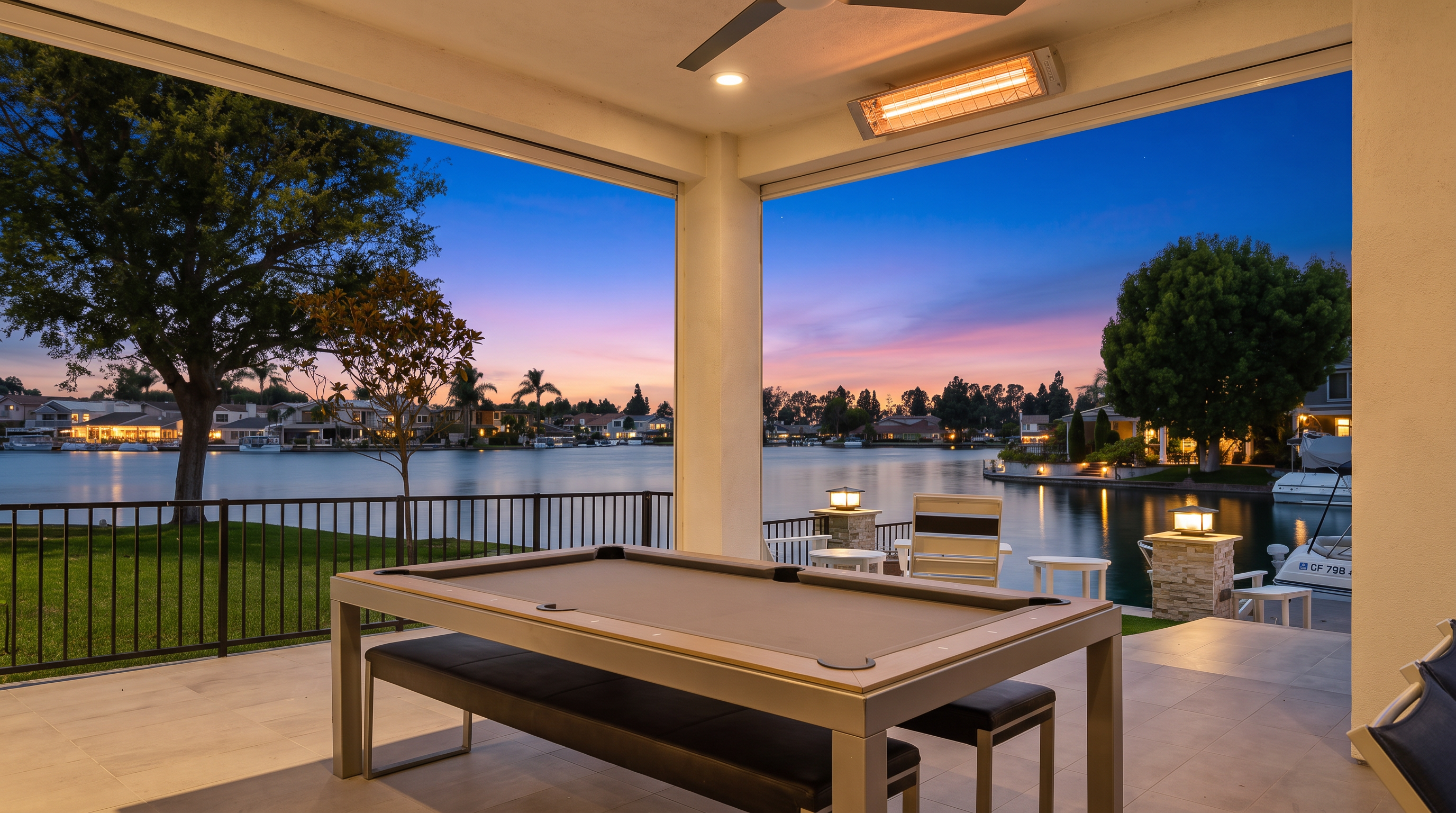 This covered outdoor patio area features a sleek, contemporary dining pool table, creating a unique space for entertainment. The space opens up directly to a stunning waterfront view, complete with a private dock and a manicured lawn. The covered ceiling is equipped with a fan and heater, making this an ideal spot for year-round outdoor enjoyment.