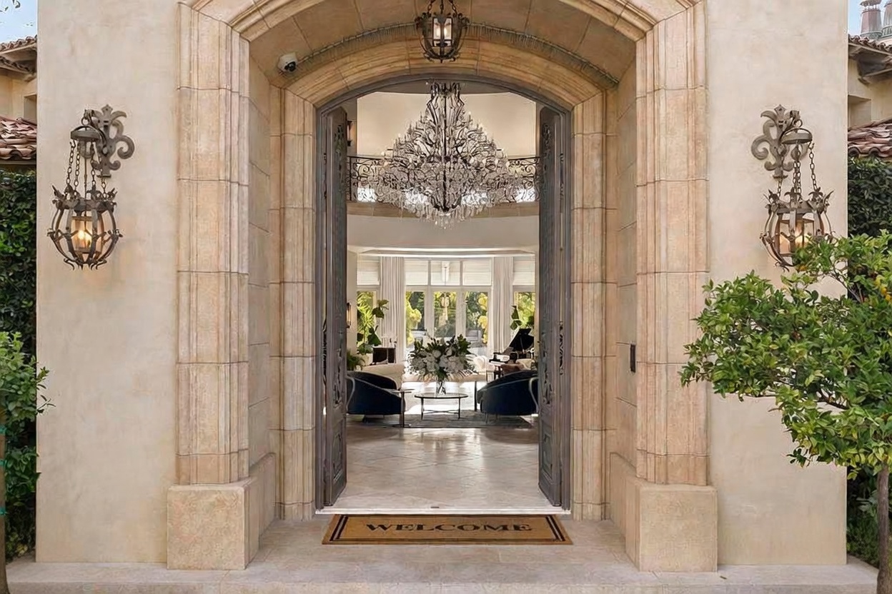 This is a grand entryway featuring an arched doorway with stone columns and ornate metal doors. A 'WELCOME' mat sits at the entrance, leading into a luxurious interior with a chandelier visible through the open doors. The exterior is adorned with decorative sconces, creating an inviting and elegant first impression.