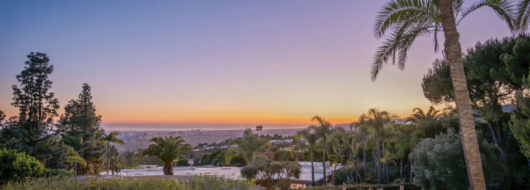 This elevated perspective captures a breathtaking sunset view overlooking a sprawling cityscape and the distant ocean horizon. The foreground features lush, mature palm trees and manicured greenery, framing the expansive vista. The scene conveys a sense of luxury and tranquility, highlighting the property's prime location and scenic outdoor environment.
