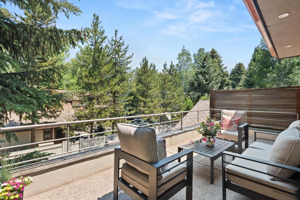 This elevated balcony offers a serene outdoor living space furnished with a modern seating set, including two armchairs and a sofa arranged around a central coffee table. The space is framed by a metal railing and a wooden privacy screen, providing a comfortable vantage point to enjoy the lush, tree-filled surroundings. The perspective captures the inviting atmosphere of the deck, highlighting its potential as a peaceful retreat for relaxation.