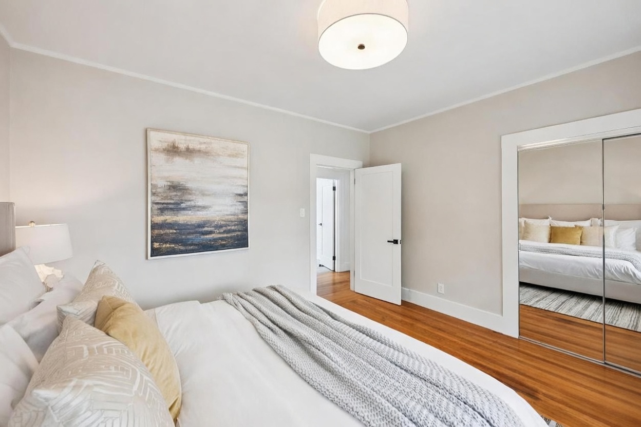This bright and airy primary bedroom features warm wood flooring and neutral wall tones, creating a serene and inviting atmosphere. A plush bed with layered bedding and a textured throw blanket serves as the focal point, complemented by a large abstract art piece on the wall and a modern drum-style ceiling light. The room includes a mirrored closet door that reflects the bed, adding depth to the space, while a small seating area with a textured armchair and side table provides a cozy corner.