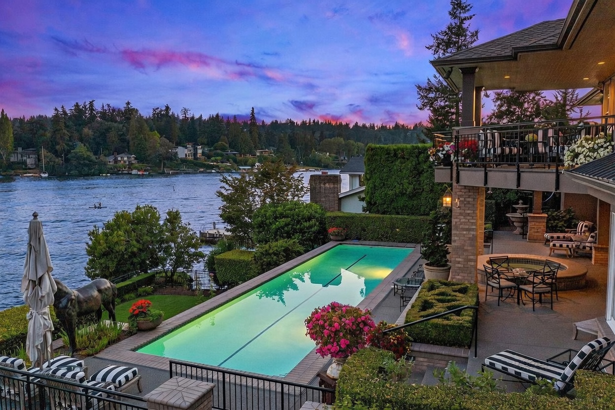 This stunning waterfront property features a long, rectangular lap pool set against a backdrop of a serene lake at twilight. The multi-level outdoor living space includes a covered patio with a dining area, a hot tub, and manicured landscaping, all framed by a dramatic purple and orange sunset. A bronze horse sculpture adds a unique artistic touch to the poolside terrace, creating an atmosphere of luxury and tranquility.