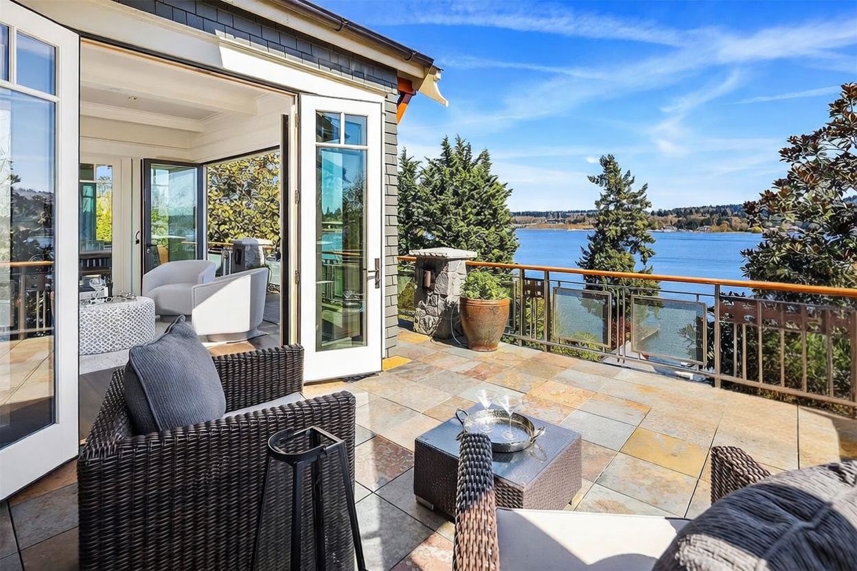 This image showcases a luxurious, elevated balcony featuring stone tile flooring and comfortable outdoor wicker furniture, perfect for relaxing while overlooking a serene lake. The space seamlessly transitions from the interior living area through large, open French doors, emphasizing an indoor-outdoor lifestyle. The scene is captured from a slightly elevated perspective, highlighting the expansive water view and the peaceful, sunny atmosphere.
