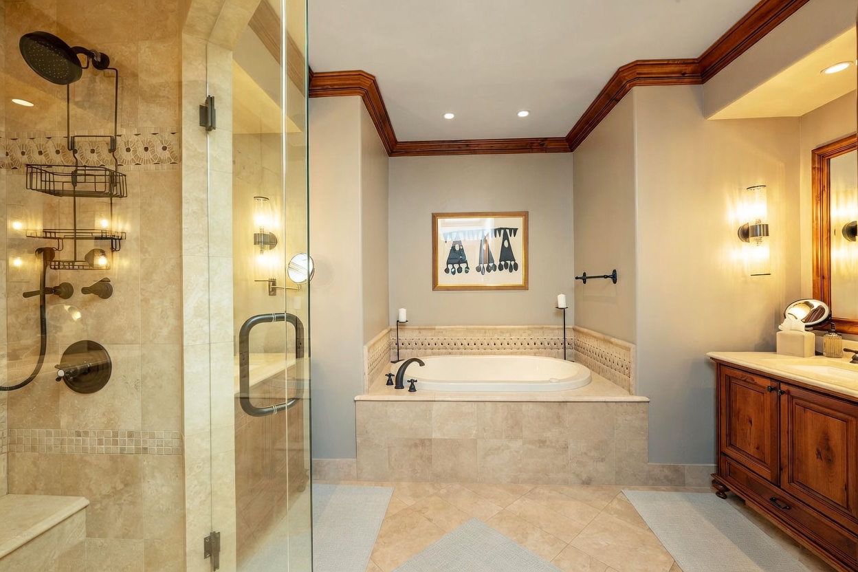 This primary bathroom features a spacious layout with a glass-enclosed walk-in shower on the left and a built-in soaking tub centered against the back wall. The room is accented by warm-toned tile flooring, a wooden vanity with a stone countertop, and crown molding that adds a traditional touch. The perspective is a wide-angle shot from the doorway, capturing the entire bathing area and the elegant, neutral color palette.
