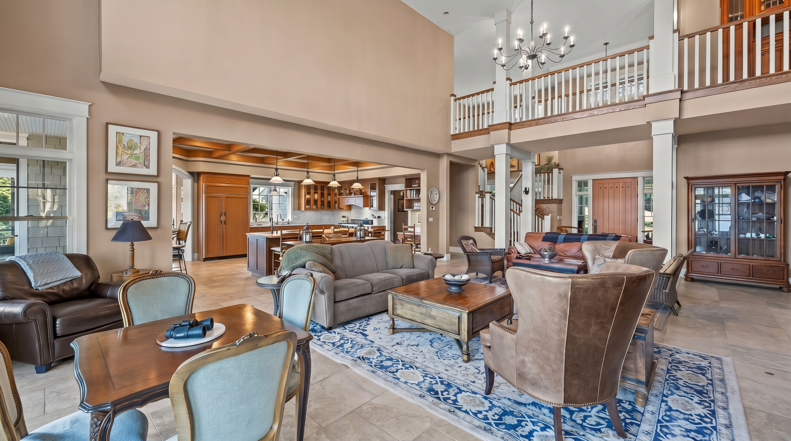 This expansive, open-concept living area features a soaring vaulted ceiling and a mezzanine level with a wooden railing overlooking the space. The room is anchored by a large patterned area rug, a leather armchair, and a central seating group, while a dining area with a wooden table sits in the foreground. In the background, a spacious kitchen with a large island and warm-toned cabinetry is visible, creating a cohesive and inviting atmosphere for entertaining.