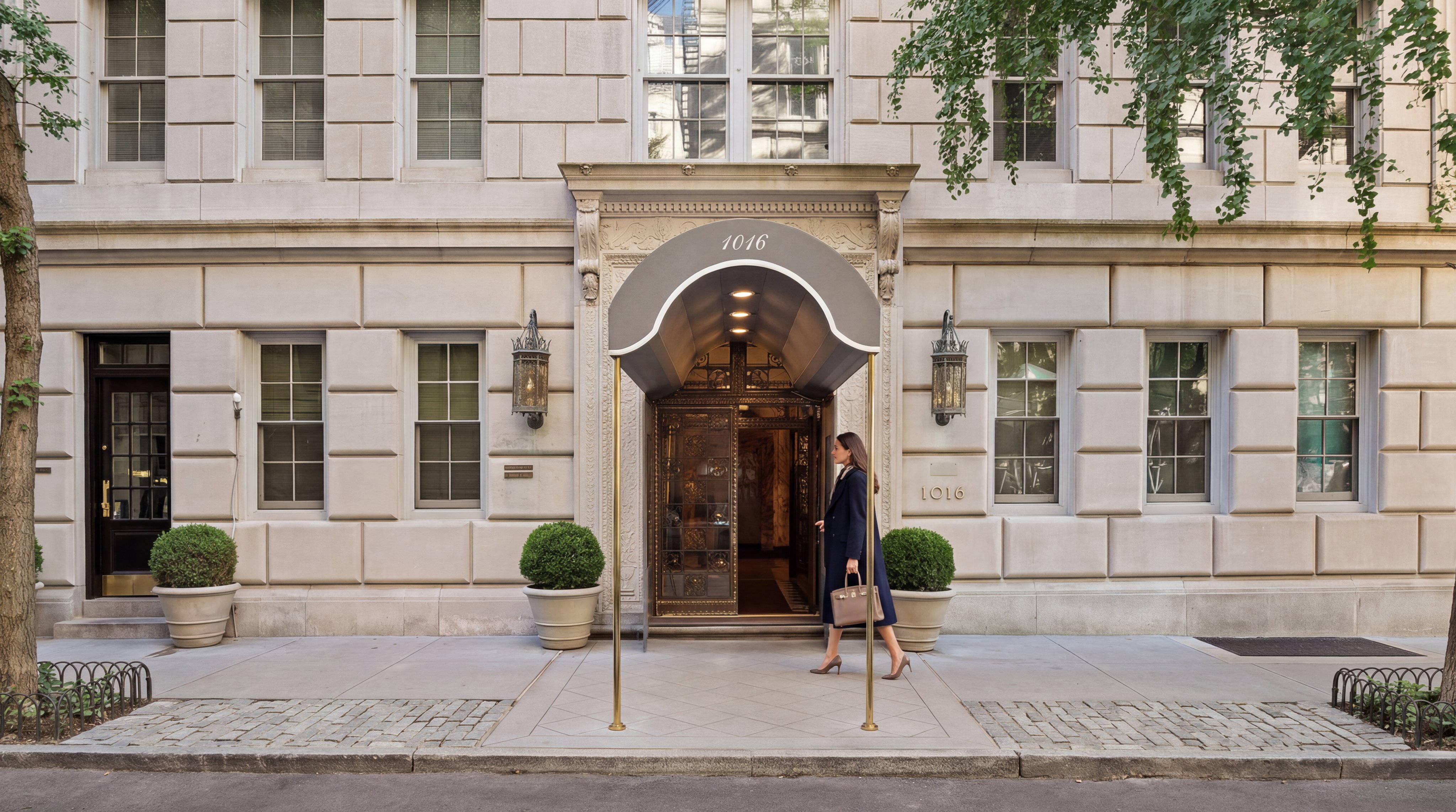 This sophisticated building entryway features a grand, classic stone facade with a decorative metal marquee marked '1016'. A woman in a professional coat is captured walking into the ornate, golden-hued entrance, which is flanked by symmetrical potted topiaries and refined lantern-style wall lights. The image conveys an air of upscale urban elegance, characteristic of a high-end pre-war residential building.