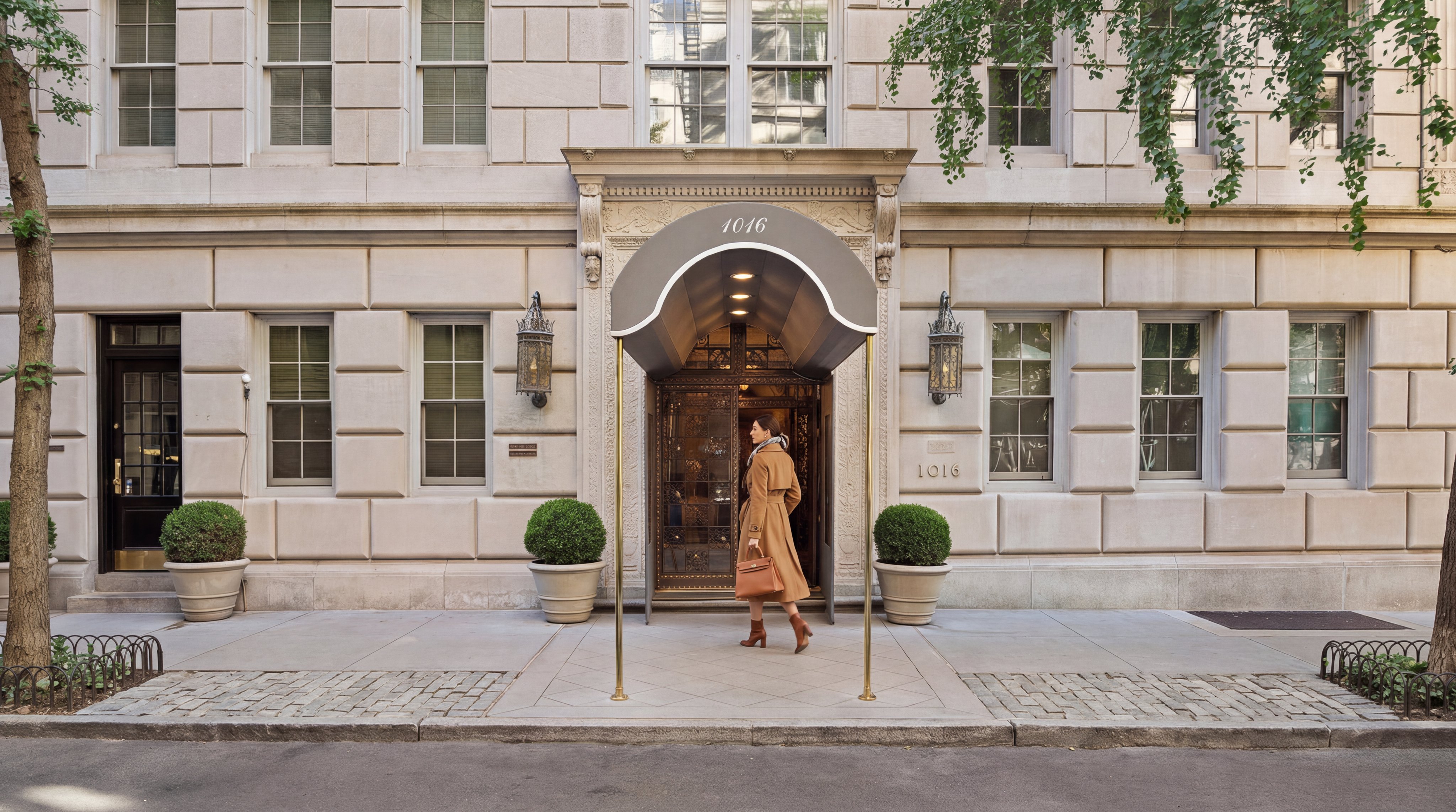 This elegant exterior entrance showcases a sophisticated limestone facade with a prominent awning marked '1016'. The symmetry of the building, featuring decorative wrought iron lamps and manicured topiaries in stone planters, projects a refined and high-end residential atmosphere. A person in a camel coat entering the building adds a sense of scale and exclusivity to this classic New York-style apartment building front.