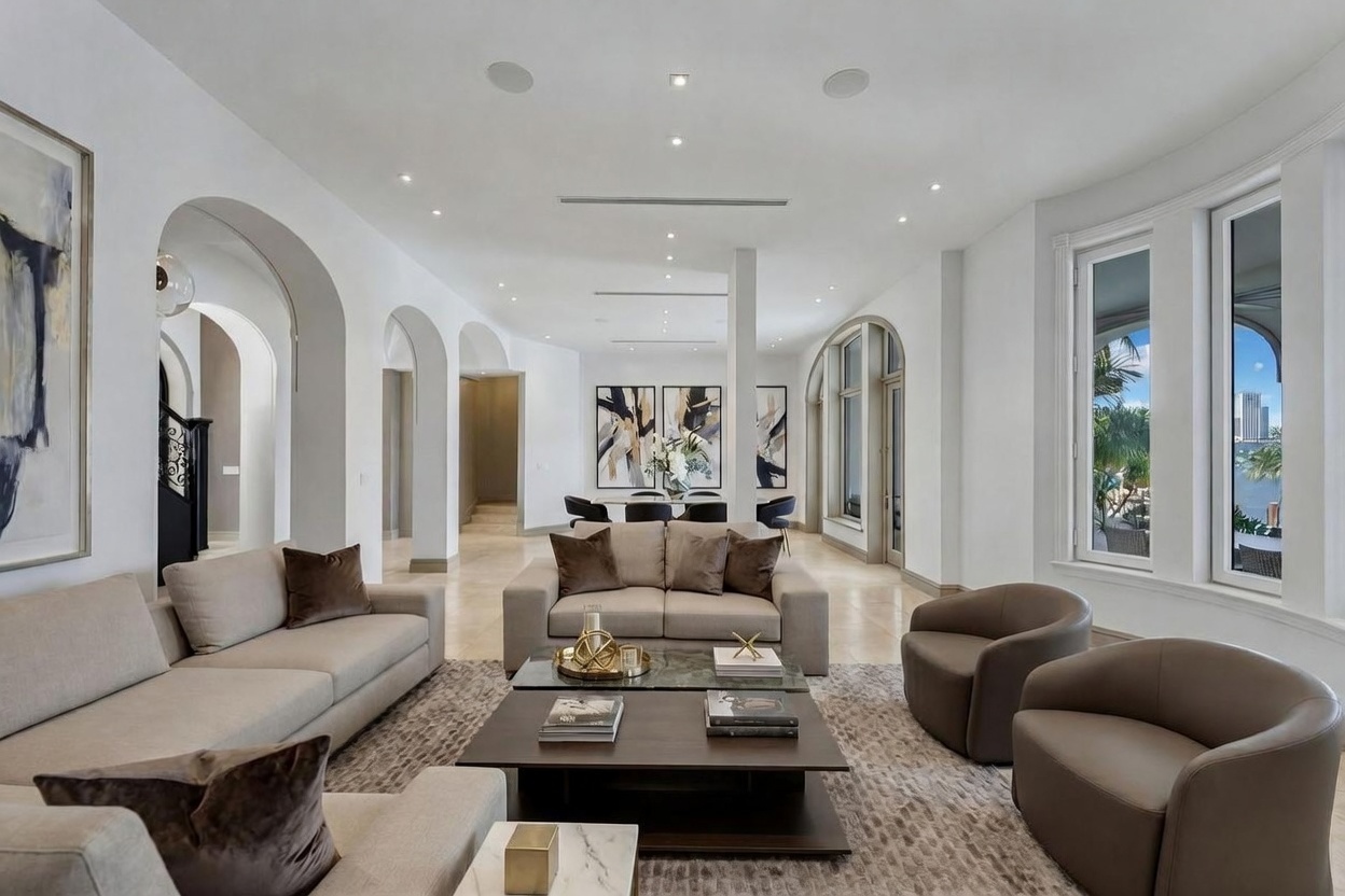 This spacious, open-concept living area features a neutral color palette with light-toned sofas and matching armchairs arranged around a tiered coffee table. The room is illuminated by recessed lighting and large windows that provide a view of the outdoors, while arched architectural openings lead to adjacent spaces. The overall aesthetic is sophisticated and contemporary, emphasizing a bright, airy atmosphere with clean lines and high-end finishes.