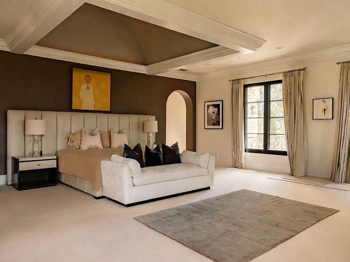 This spacious primary bedroom features a large, upholstered headboard against a dark accent wall, complemented by a matching bench at the foot of the bed. The room is anchored by a neutral-toned area rug and features a tray ceiling with recessed lighting, creating an elegant and serene atmosphere. Large windows with floor-length curtains allow natural light to fill the space, while an arched doorway adds a touch of architectural interest.