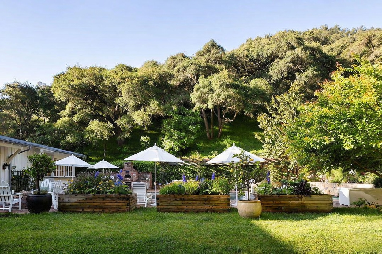 This lush backyard features a well-manicured lawn leading to a series of raised wooden garden beds filled with vibrant flowers and greenery. Several white umbrellas provide shade over the patio area, which includes a brick outdoor fireplace structure nestled against a backdrop of dense, mature trees on a hillside. The scene captures a peaceful, private outdoor living space perfect for relaxation or entertaining.