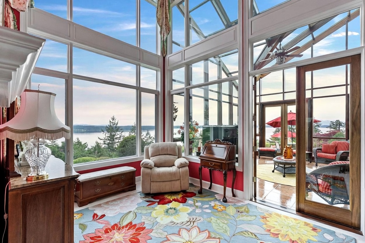 This bright sunroom features expansive glass walls that offer panoramic views of the water and surrounding trees. The space is furnished with a comfortable armchair, a small wooden writing desk, and a patterned area rug, creating a cozy reading nook. A sliding glass door leads to an adjacent covered patio, seamlessly blending indoor and outdoor living areas.