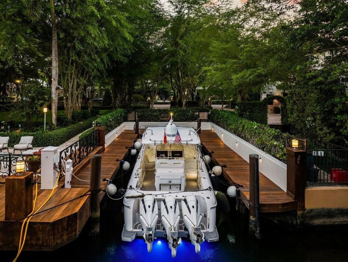 This elevated perspective captures a private boat slip nestled within a lush, landscaped canal-side setting. A sleek white motorboat with triple outboard engines is docked between wooden piers, illuminated by vibrant blue underwater lights. The scene is framed by mature trees and manicured hedges, creating a serene and exclusive waterfront atmosphere.