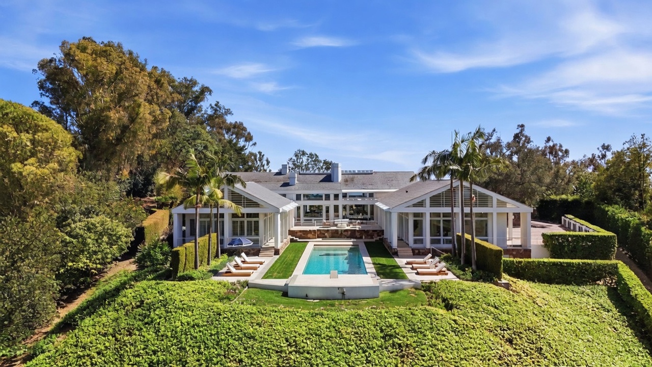 This expansive rear view captures a luxurious estate featuring a symmetrical white architectural design with large windows and multiple glass doors opening to the backyard. A rectangular swimming pool serves as the centerpiece of the manicured lawn, flanked by lounge chairs and tall palm trees. The property is surrounded by lush, mature landscaping and dense greenery, creating a private and serene atmosphere.