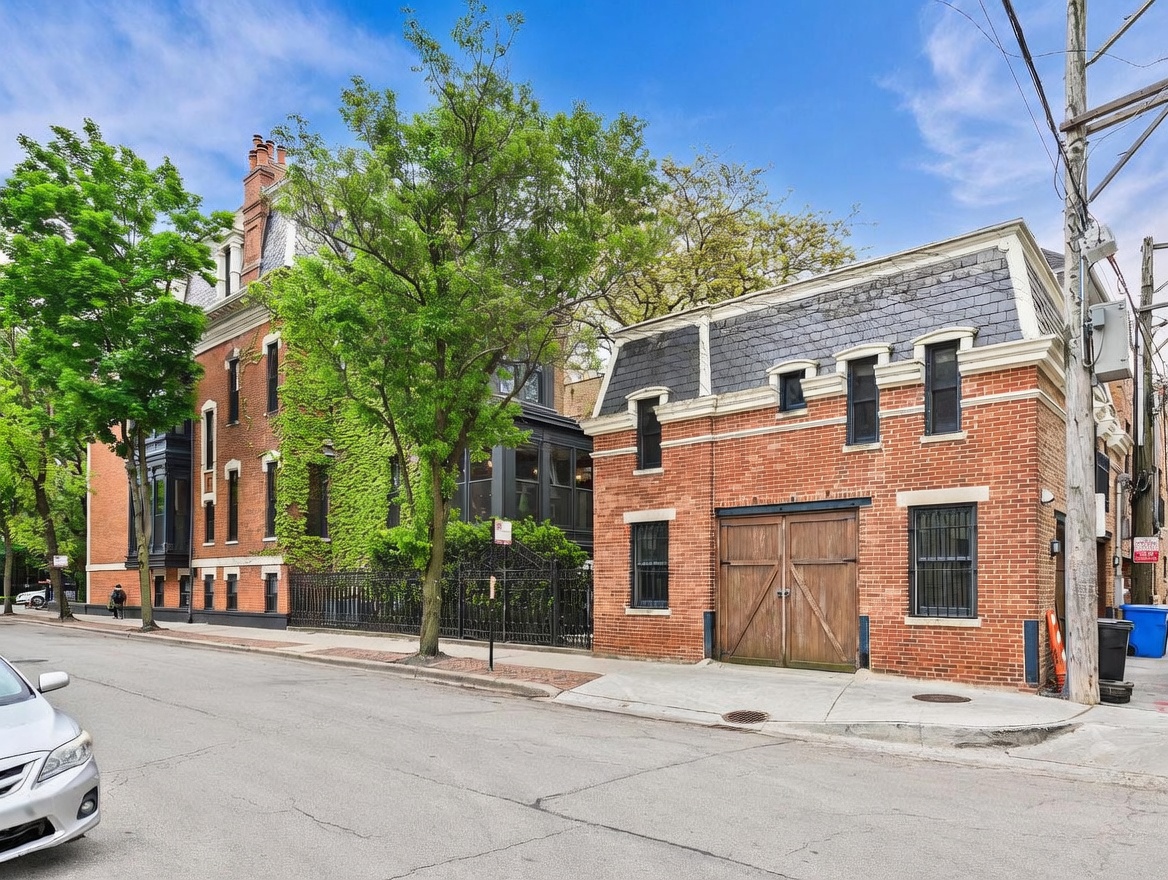 This street-level view captures a charming urban scene featuring a historic brick building adorned with climbing ivy alongside a separate, smaller brick carriage house. The carriage house showcases a classic mansard roofline and large wooden double doors, suggesting a converted garage or studio space. The perspective is from the street, highlighting the architectural character and the mature trees that frame the properties.