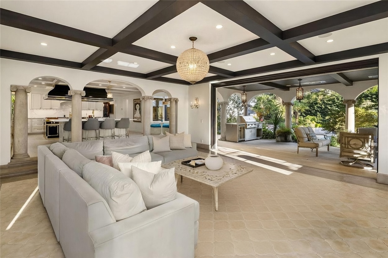 This expansive living room features a coffered ceiling with dark beams and a large crystal chandelier, seamlessly transitioning to an outdoor patio through a wide, open-wall design. The space is furnished with a large light-colored sectional sofa, a patterned area rug, and a central coffee table, while stone columns frame the view into the adjacent kitchen and the outdoor living area. The overall aesthetic is one of luxury and indoor-outdoor integration, perfect for entertaining.