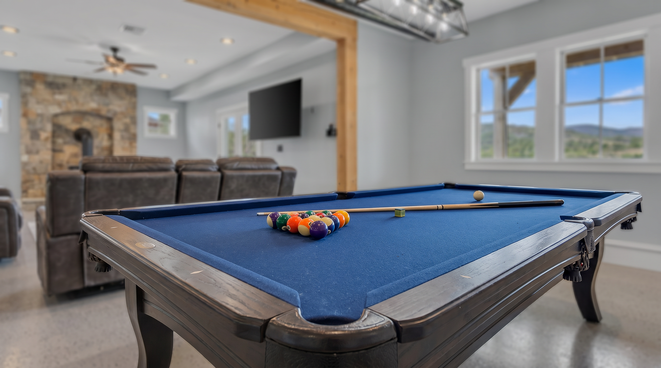 This spacious, open-concept basement features a dedicated game area with a blue-felt pool table and a cozy living space anchored by a stone-clad chimney breast with a wood-burning stove. The room is defined by a prominent exposed timber beam that separates the two zones, while large windows provide natural light and scenic views of the surrounding landscape. The polished concrete flooring and neutral wall tones create a versatile, modern recreational environment.
