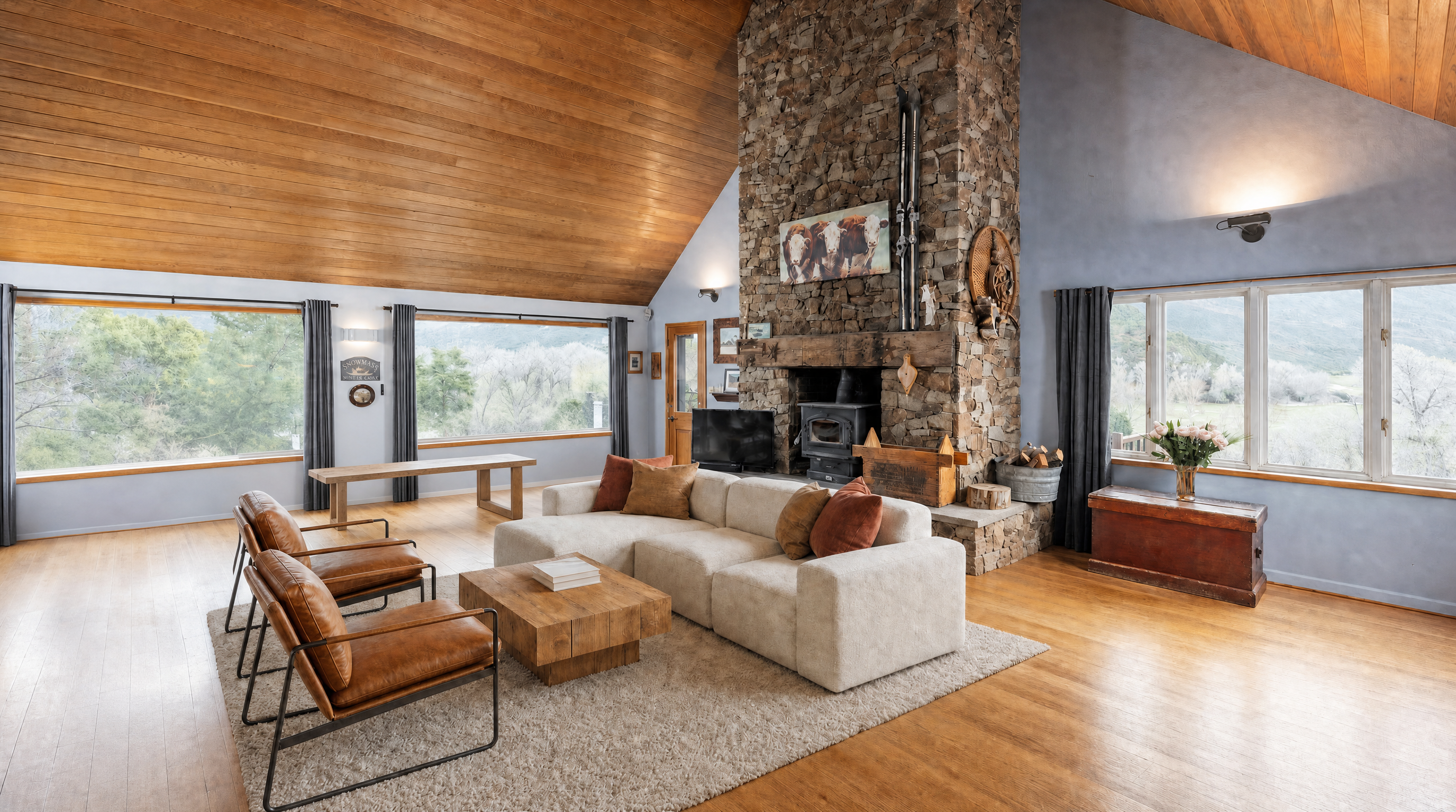 This spacious and airy living room features a vaulted pine ceiling and a dramatic floor-to-ceiling stone fireplace with a wood-burning stove. The open-concept design flows seamlessly into the dining area, accented by warm hardwood floors and large windows that offer expansive views of the surrounding natural landscape. The room is decorated in a comfortable, rustic-modern style, highlighted by leather accent chairs, a light-toned sectional, and natural wood furniture pieces.