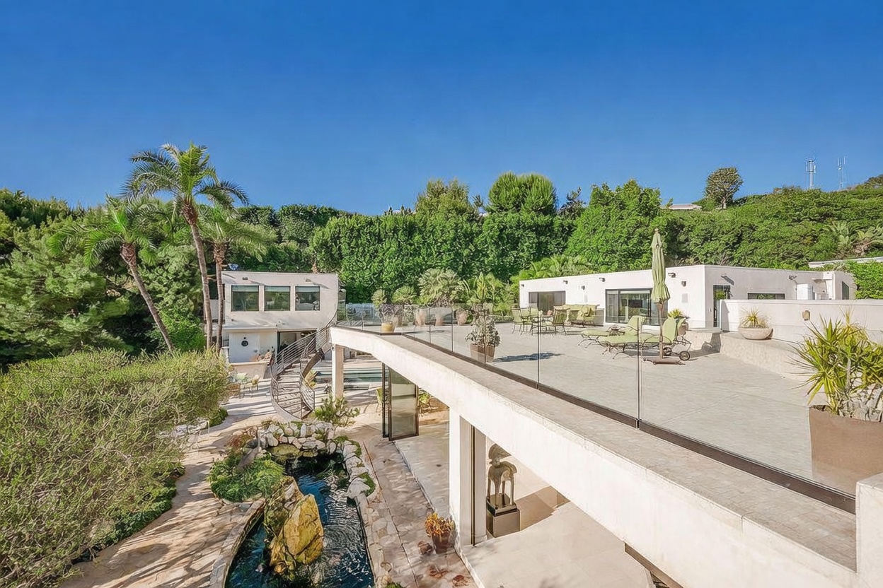 This rear view of a modern residence showcases a multi-level architectural design with a spacious rooftop terrace enclosed by glass railings. Below the terrace, a tranquil koi pond with large decorative rocks sits adjacent to a covered patio area, creating a seamless transition between indoor and outdoor living. The property is nestled against a lush, green hillside, emphasizing a private and serene atmosphere.