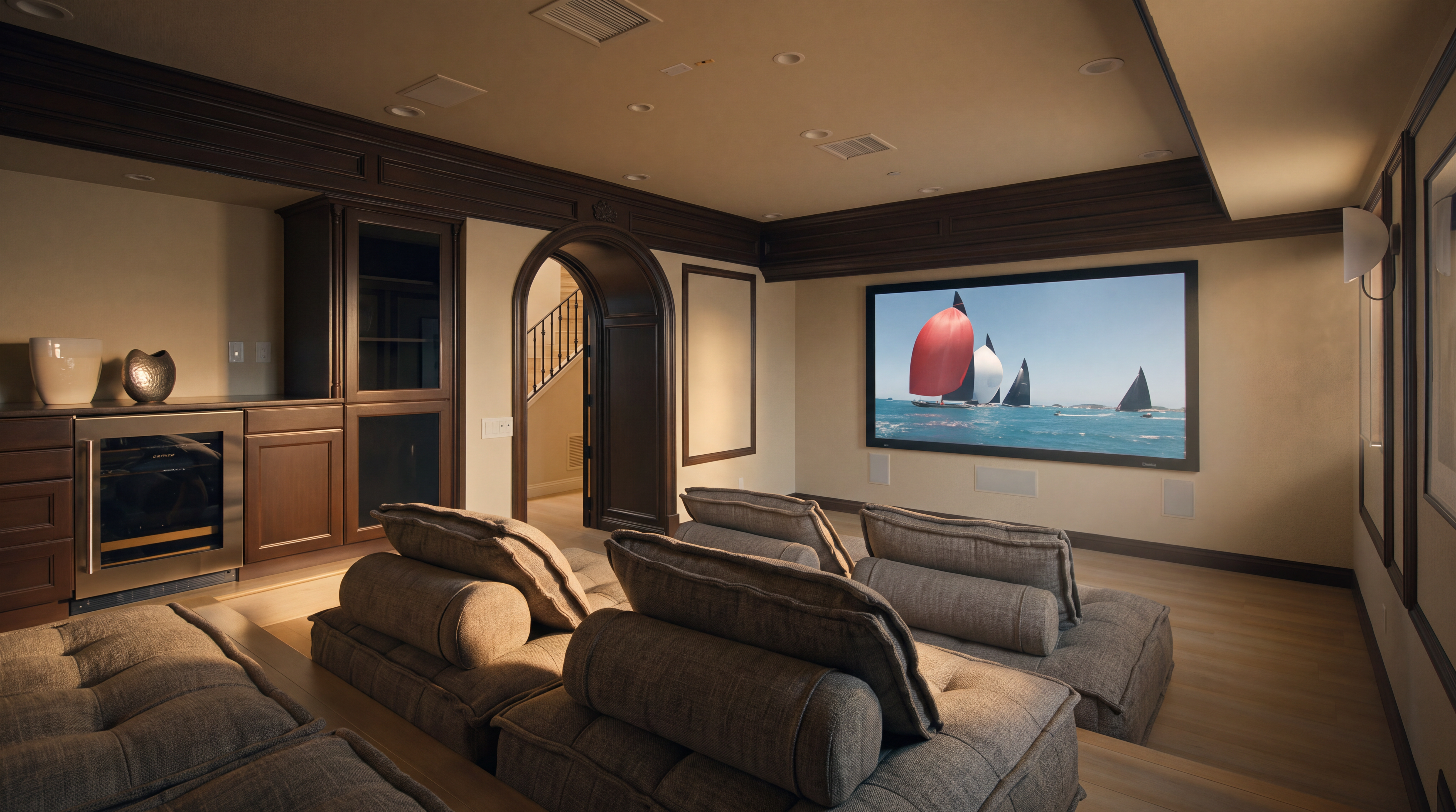 This dedicated home theater room features tiered seating with plush, grey modular sofas arranged for optimal viewing of a large wall-mounted screen. The space is accented by dark wood crown molding and trim, a built-in wet bar with a wine refrigerator, and an arched doorway leading to a staircase. The room is illuminated by recessed ceiling lights, creating a sophisticated and comfortable environment for entertainment.