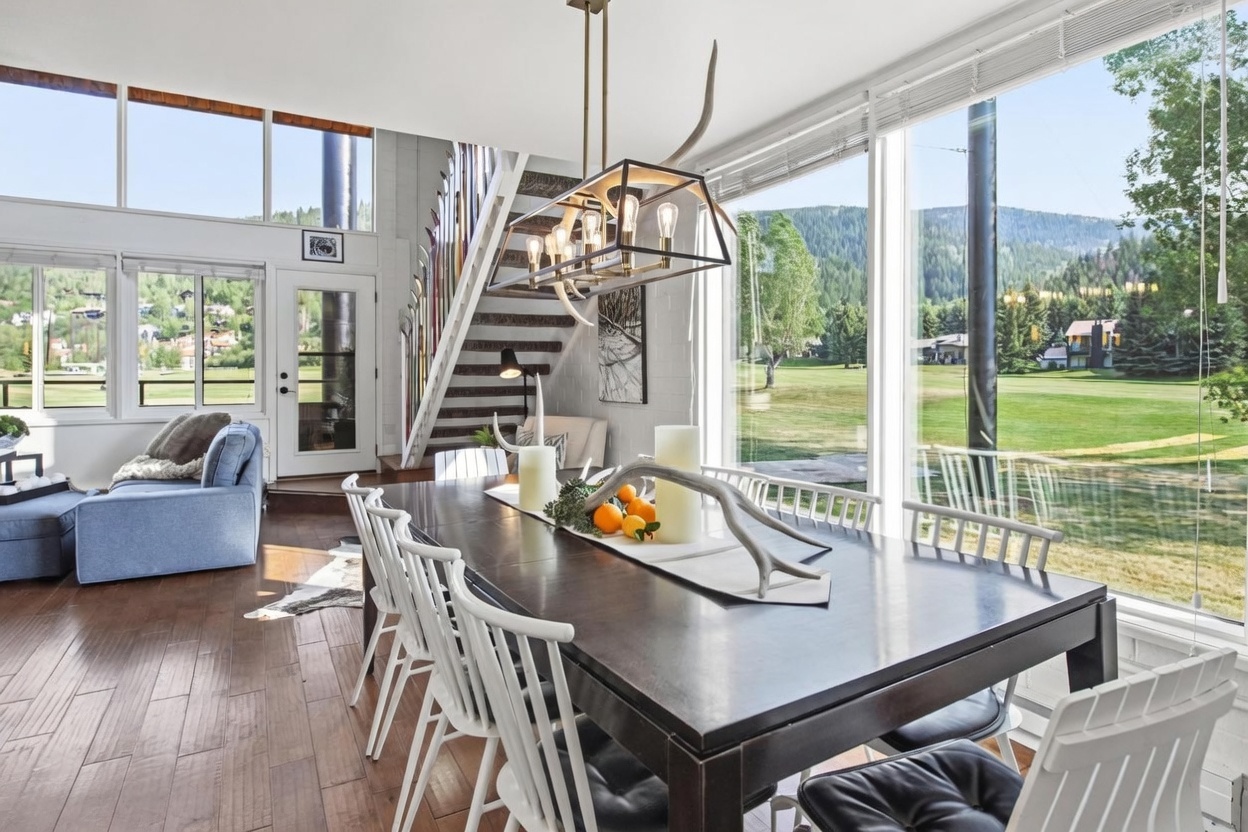 This bright dining area features a dark wood table surrounded by white spindle-back chairs, set against a backdrop of expansive windows that offer panoramic views of a lush green landscape. A unique antler-accented chandelier hangs above the table, which is decorated with a centerpiece of candles and fresh citrus. The space flows seamlessly into an adjacent living area with a blue sofa and a staircase lined with decorative skis, creating a cozy, mountain-inspired atmosphere.