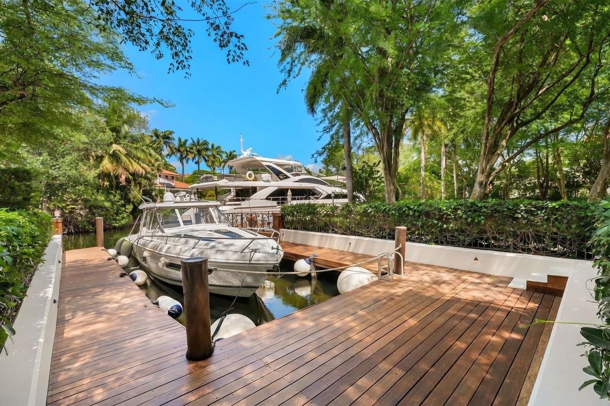 This image captures a private wooden boat dock extending over calm water, featuring a sleek motorboat moored alongside. The dock is framed by lush green trees and a manicured hedge, creating a serene and secluded waterfront atmosphere. In the background, a larger yacht is visible, emphasizing the property's prime location for boating enthusiasts.