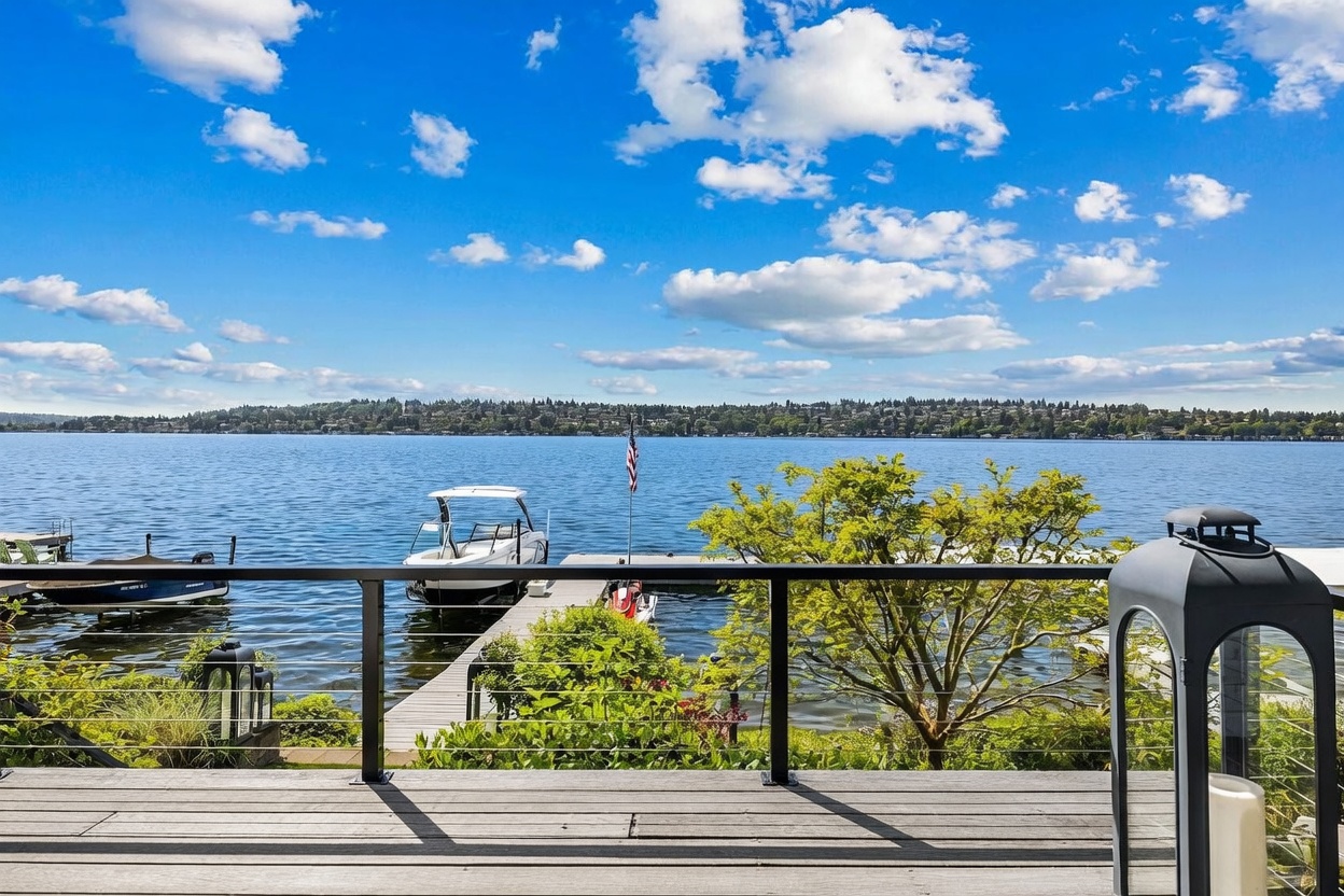 This elevated deck offers a stunning, unobstructed view of a large lake under a bright blue sky filled with scattered clouds. The foreground features a modern railing with horizontal cables, while the mid-ground showcases a private dock with a boat and a personal watercraft. Lush green trees and foliage frame the view, creating a serene and picturesque waterfront setting.