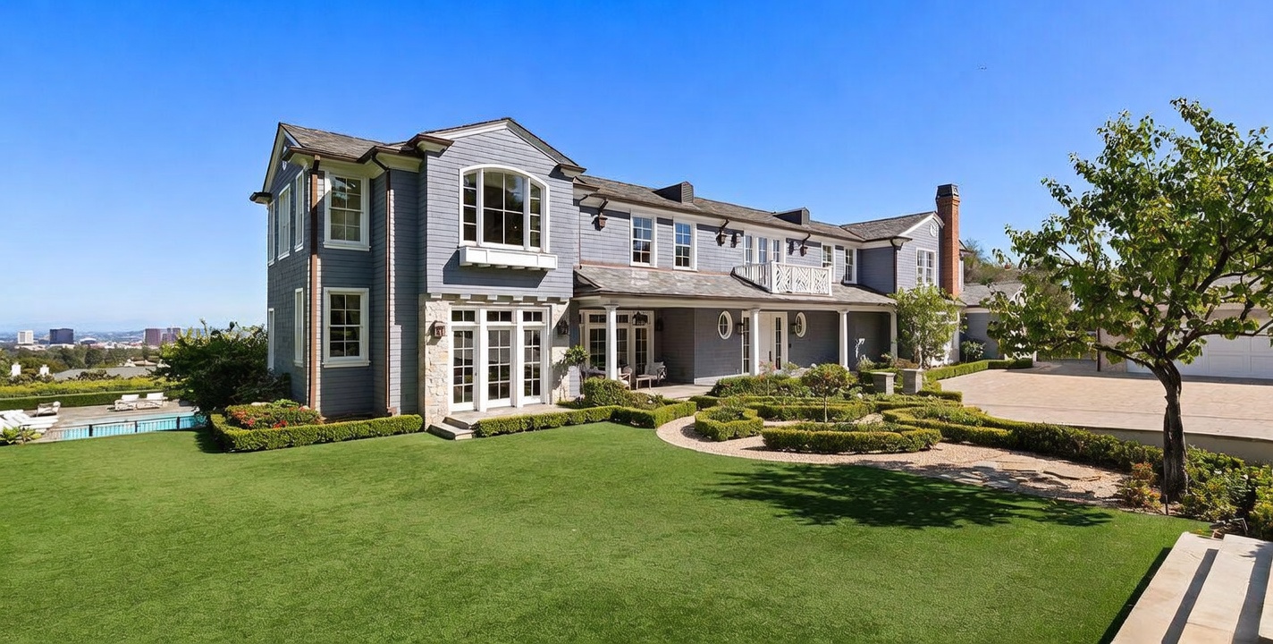 This expansive rear view of a multi-story traditional home showcases a classic gray shingle exterior with stone accents and a sprawling, manicured lawn. The property features a formal garden with neatly trimmed hedges, a paved driveway area, and a glimpse of a swimming pool to the left. The composition captures the home's architectural elegance against a clear blue sky, highlighting its integration with the landscaped grounds and distant city views.