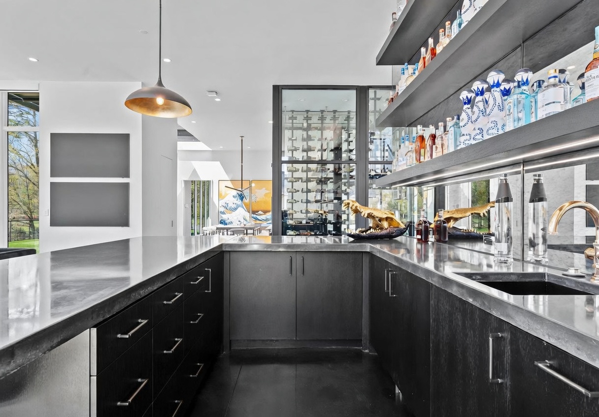 This sophisticated home bar area features dark, sleek cabinetry paired with a metallic countertop and a mirrored backsplash that reflects the room. A glass-enclosed wine display serves as a striking focal point, while open shelving displays an extensive collection of spirits. The space is illuminated by a modern pendant light, creating a moody and upscale atmosphere perfect for entertaining.