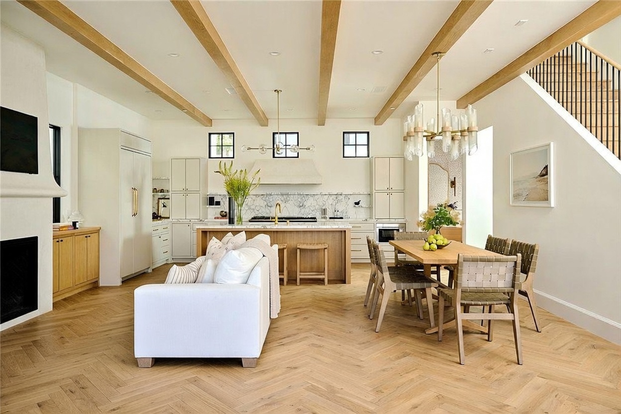 This open-concept living and dining area features a bright, modern kitchen with light wood cabinetry, a central island, and a stone-topped breakfast bar. The space is defined by light wood herringbone flooring and exposed ceiling beams, creating a cohesive and airy atmosphere. A dining table sits adjacent to the kitchen, while a comfortable living area with a fireplace is positioned in the foreground, offering a seamless flow for entertaining.