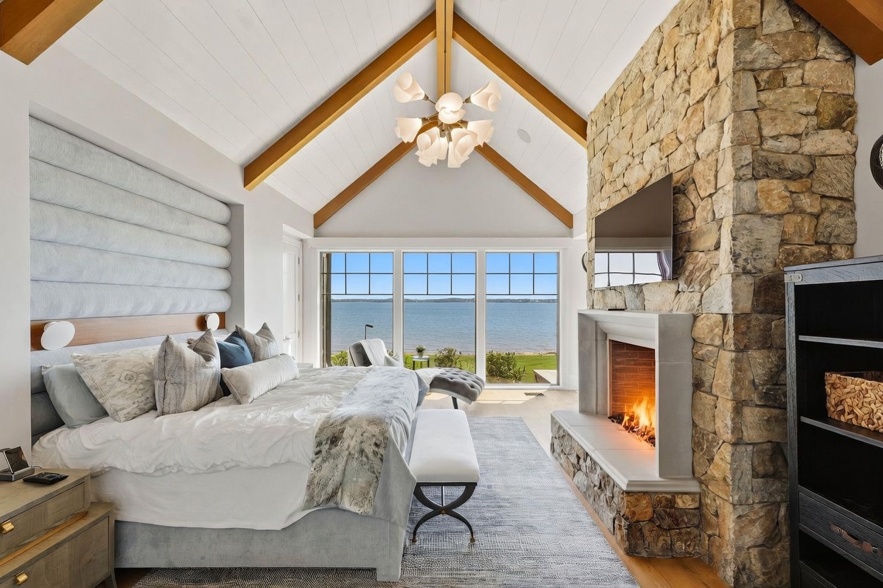 This luxurious primary bedroom features a vaulted ceiling with exposed light wood beams and a striking stone-clad chimney breast housing a fireplace. A large, plush bed sits against a textured, upholstered wall panel, while expansive windows offer a panoramic view of the water. The room is anchored by a soft area rug and illuminated by a modern, multi-armed chandelier, creating a serene and sophisticated atmosphere.
